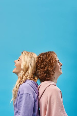 Photo for Side view of excited blonde girl with redhead friend laughing with closed eyes back to back on blue - Royalty Free Image