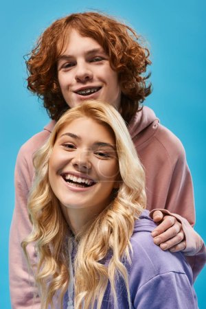 portrait of carefree teenage friends, redhead guy and blonde girlfriend looking at camera on blue
