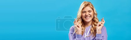 worried blonde teen girl with closed eyes holding crossed fingers for luck on blue, banner