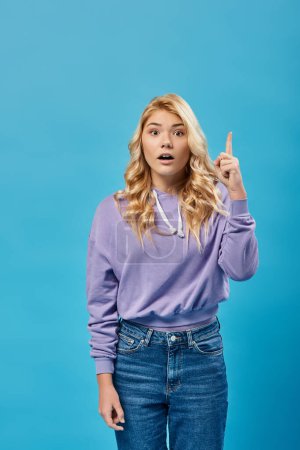 inspirierte blonde Teenager-Mädchen mit offenem Mund zeigt Idee Geste und Blick in die Kamera auf blau