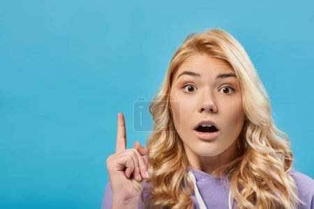 Porträt eines erstaunten blonden Teenagermädchens mit offenem Mund, das Ideengeste auf blau zeigt