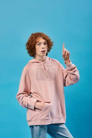 amazed redhead teen guy showing idea sign, holding hand in pocket and looking at camera on blue