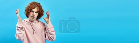 Téléchargez les photos : Adolescent en colère avec les cheveux roux ondulés regardant la caméra et gesticulant sur bleu, mauvaise humeur, bannière - en image libre de droit