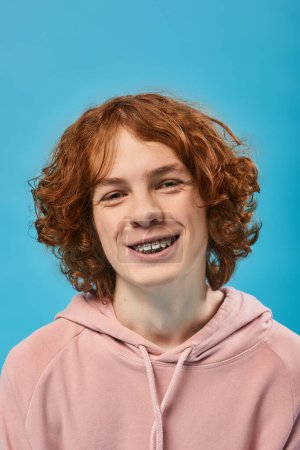 Téléchargez les photos : Portrait d'adolescent gai avec des cheveux roux ondulés et des bretelles souriant à la caméra sur bleu, bonheur - en image libre de droit