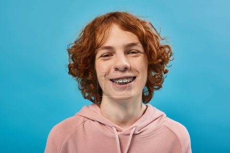 Téléchargez les photos : Adolescent insouciant et joyeux avec des cheveux roux ondulés et des accolades regardant la caméra sur bleu, portrait - en image libre de droit