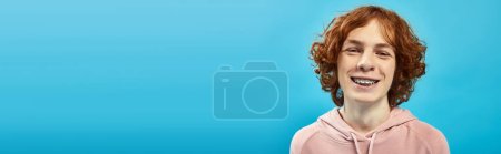 Téléchargez les photos : Portrait d'adolescent joyeux avec des cheveux roux ondulés et des bretelles souriantes à la caméra sur bleu, bannière - en image libre de droit