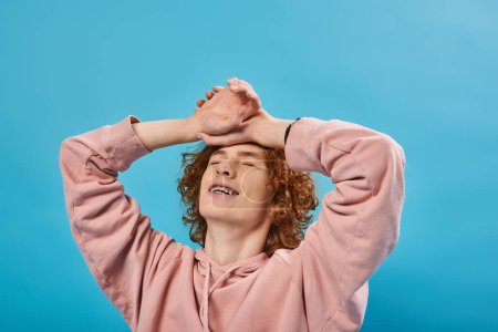 Téléchargez les photos : Positif, rêveur rousse adolescent gars en sweat à capuche debout avec les yeux fermés et les mains sur le front sur le bleu - en image libre de droit