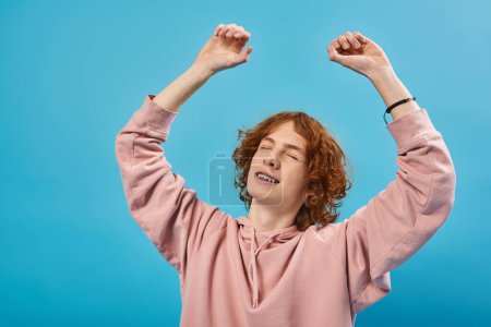 Téléchargez les photos : Sourire et rêveur rousse adolescent gars en sweat à capuche debout avec les yeux fermés et les mains levées sur le bleu - en image libre de droit