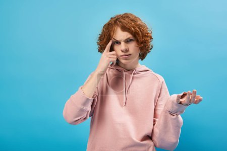 Téléchargez les photos : Réfléchi rousse adolescent gars à la mode sweat à capuche toucher la tête, geste et regarder la caméra sur bleu - en image libre de droit