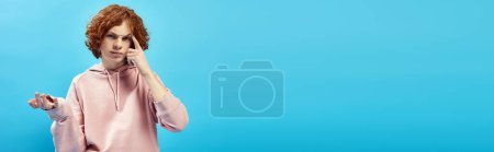 Téléchargez les photos : Onéreux rousse adolescent garçon à la mode sweat à capuche toucher la tête et regarder la caméra sur bleu, bannière - en image libre de droit