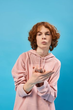 Téléchargez les photos : Adolescent sérieux avec des cheveux roux ondulés portant un sweat à capuche élégant et regardant caméra sur bleu - en image libre de droit