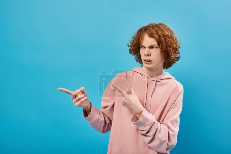 Téléchargez les photos : Mécontent rousse adolescent garçon en sweat à capuche à la mode regardant loin et pointant avec les doigts sur bleu - en image libre de droit