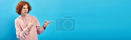 Téléchargez les photos : Rousse irritée adolescent garçon en sweat à capuche à la mode regardant loin et pointant avec les doigts sur bleu, bannière - en image libre de droit