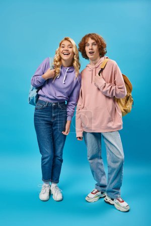 Foto de Alegre adolescente estudiantes en moda sudaderas con capucha y vaqueros posando con mochilas en azul, longitud completa - Imagen libre de derechos