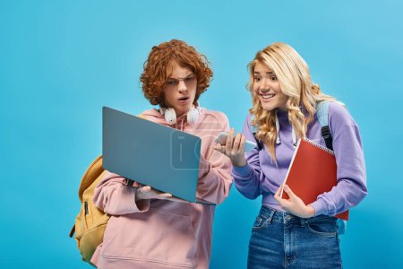 excited teen girl with smartphone pointing at laptop near thoughtful redhead student on blue