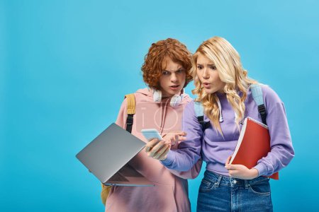 estudiantes adolescentes asombrados con mochilas, ordenador portátil y portátil mirando el teléfono inteligente en blues