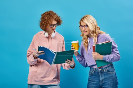 thoughtful redhead teen student showing notebook to blonde girlfriend with paper cup on blue