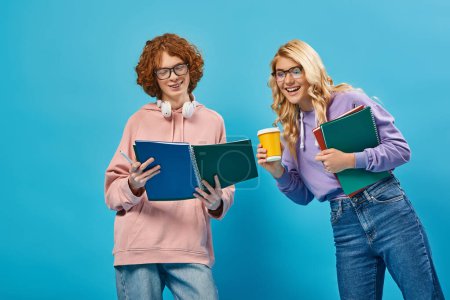cheerful blonde teen girl with paper cup laughing near redhead student with notebook on blue