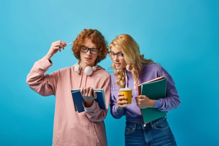 thoughtful redhead student in eyeglasses holding notebooks near girlfriend with paper cup on blue