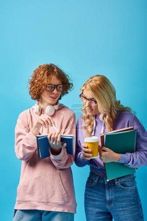 smiling redhead student in eyeglasses pointing at notebook near teen girl with coffee to go on blue