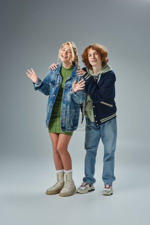rothaarige Teenager-Kerl umarmt Schultern der freudigen blonden Freundin auf grau, Mode und Glück