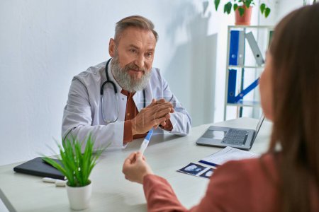 serious grey bearded doctor looking carefully at female patient showing pregnancy test, ivf concept