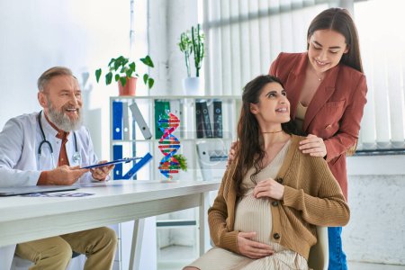 cheerful grey bearded doctor looking happily at loving lesbian couple with hands on pregnant belly