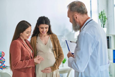 cheerful lesbian couple at doctor checkup looking at pregnant belly, in vitro fertilization concept