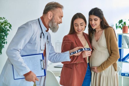 grey bearded doctor showing ultrasound to happy lesbian married couple, in vitro fertilizing concept