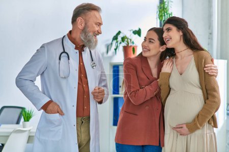 happy lesbian couple hugging with hands on pregnant belly and looking at their doctor, ivf concept