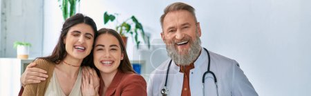grey bearded cheerful doctor smiling at camera standing near smiley lgbt couple, ivf concept, banner
