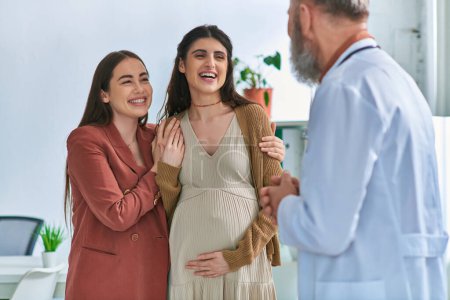 freudiges lesbisches Paar mit Händen auf schwangerem Bauch, das ihren Frauenarzt anlächelt, ivf concept