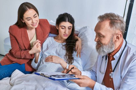 cheerful grey bearded doctor showing ultrasound to lesbian couple, in vitro fertilization concept