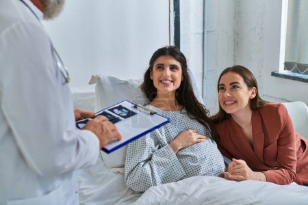 cheerful lesbian couple smiling at doctor holding ultrasound of baby, in vitro fertilization concept