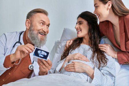Photo for Smiley grey bearded doctor showing ultrasound to hugging lgbt couple looking at them, ivf concept - Royalty Free Image