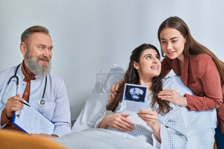 happy lesbian couple hugging and looking at ultrasound with doctor next to them, ivf concept