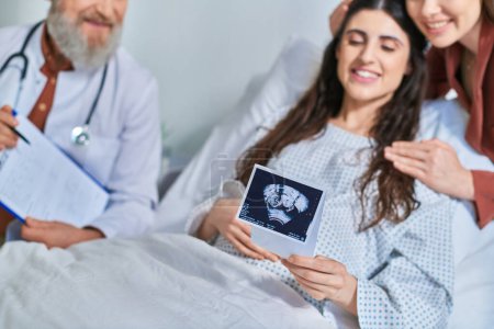 cropped view of happy lesbian couple looking at ultrasound with doctor next to them, ivf concept