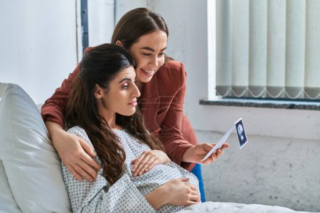 young lesbian couple looking at ultrasound of their baby with hands on pregnant belly, ivf concept