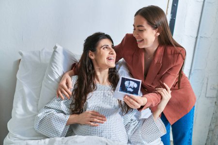 smiley beautiful lgbt couple looking at each other holding ultrasound of their baby, ivf concept