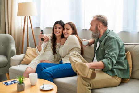 glücklicher Vater auf dem Sofa mit seiner Tochter und ihrem Partner beim Selfie-Kaffeetrinken, ivf concept