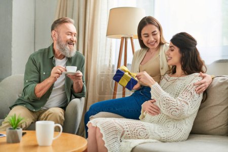 cheerful lesbian couple holding present and father of one of them drinking coffee, ivf concept