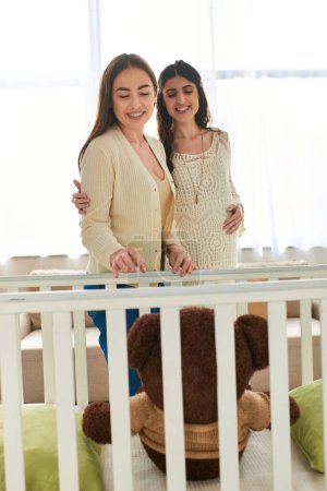 fröhliches lesbisches Paar, lächelnd und mit Blick auf einen riesigen Teddybär in der Krippe, Hand auf Bauch, ivf concept