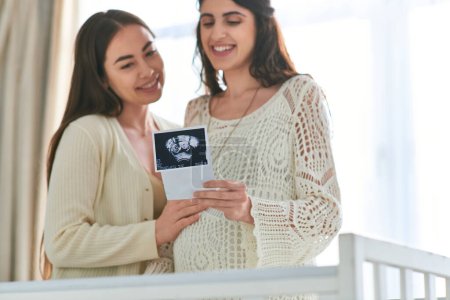 cheerful lgbt couple standing next to crib looking at ultrasound of their baby, ivf concept