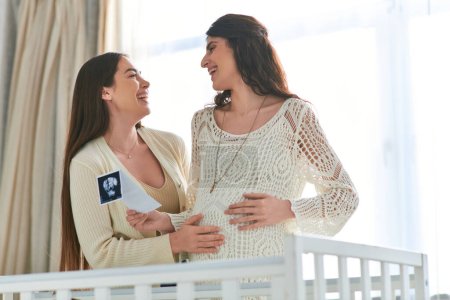 happy lesbian couple near crib holding ultrasound and smiling sincerely at each other, ivf concept