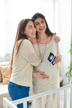 cheerful lesbian couple near crib hugging warmly and smiling with ultrasound in hands, ivf concept