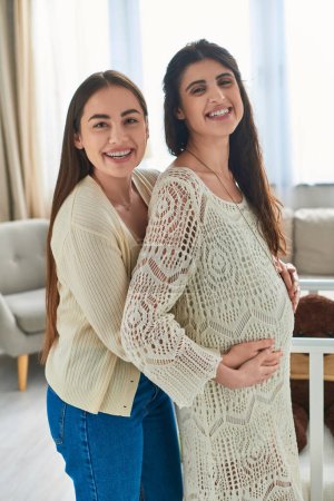 cheerful lesbian couple hugging with hands on pregnant belly smiling at camera, ivf concept