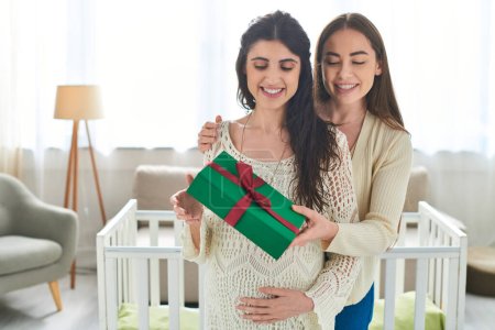 pretty lesbian couple standing next to crib holding present with hand on pregnant belly, ivf concept