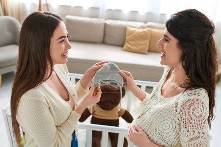 cheerful lgbt couple holding beanie and smiling at each other with crib on backdrop, ivf concept