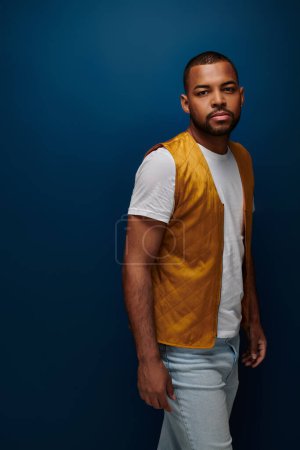 hombre afroamericano de buen aspecto en chaleco amarillo y jeans sobre fondo azul oscuro, concepto de moda