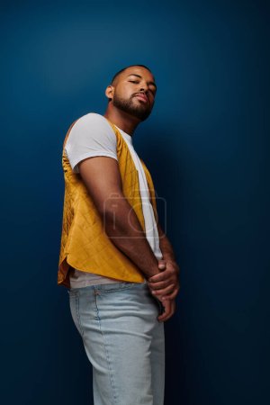 Téléchargez les photos : Élégant homme afro-américain en gilet jaune posant de profil et regardant la caméra, concept de mode - en image libre de droit